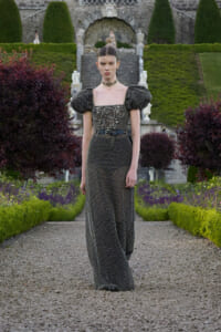 Model wearing a dark, beaded gown with puff sleeves and a choker in a formal garden setting.