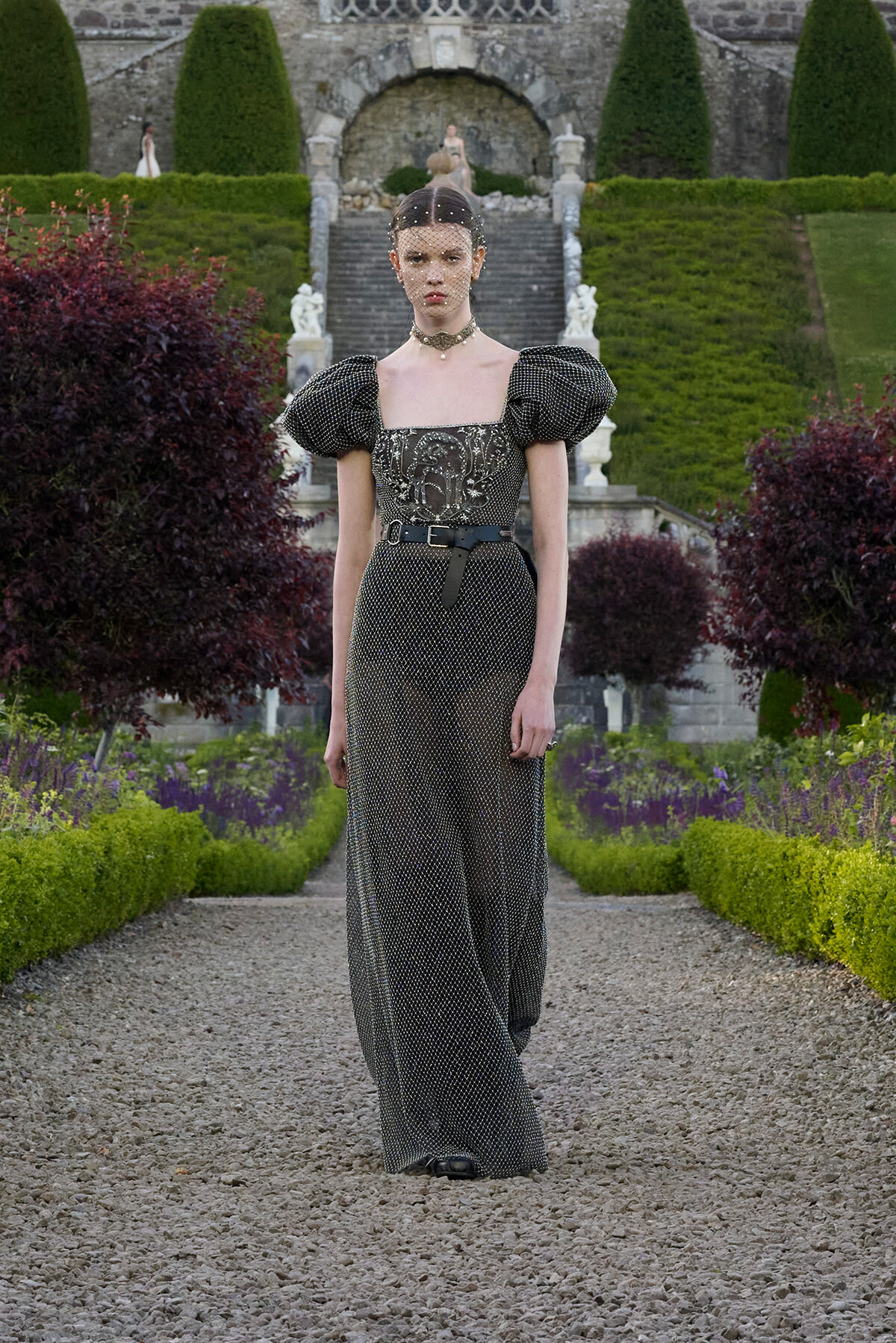 Model wearing a dark, beaded gown with puff sleeves and a choker in a formal garden setting.