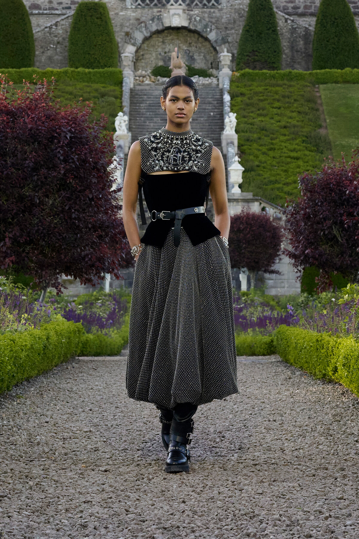 Model posing in a formal garden wearing a black embellished top, wide dotted skirt, and chunky boots.