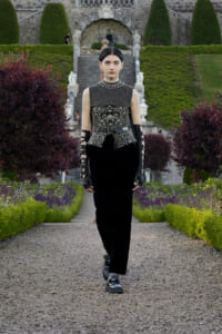 Model in a black embellished sleeveless top and wide belt walking toward the camera along a gravel garden path, stone stairs in background.
