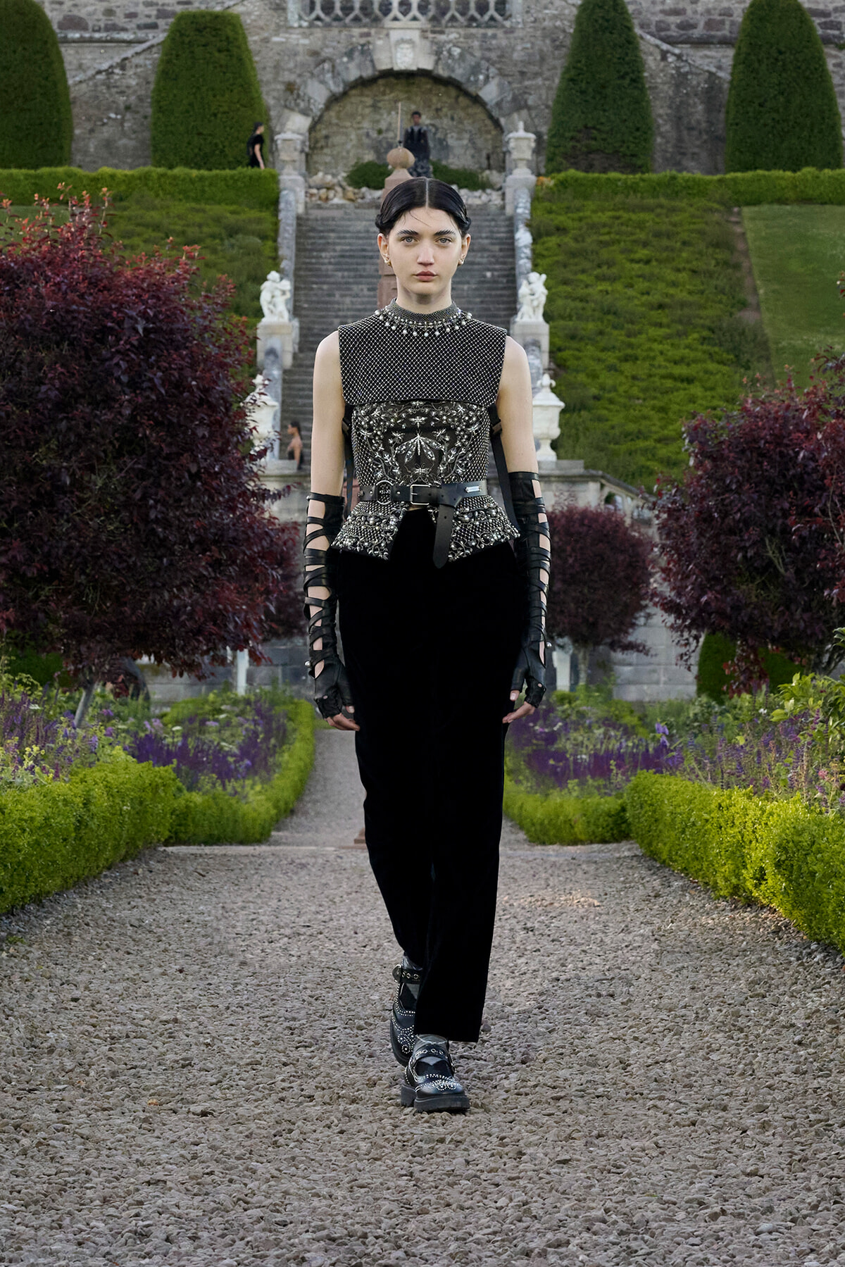 Model in a black embellished sleeveless top and wide belt walking toward the camera along a gravel garden path, stone stairs in background.