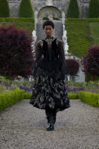 Person wearing a black embroidered Victorian-style gown and boots walking along a gravel garden path in a formal garden setting.