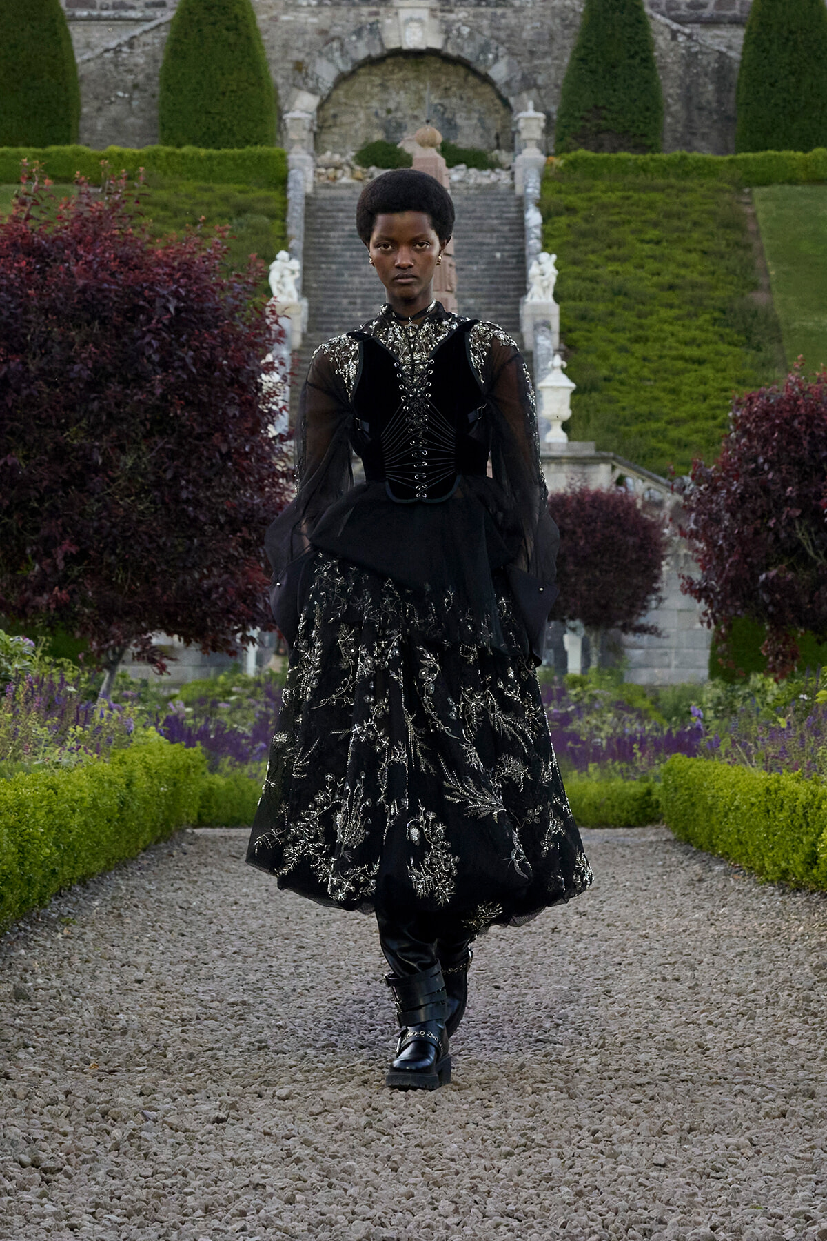Person wearing a black embroidered Victorian-style gown and boots walking along a gravel garden path in a formal garden setting.