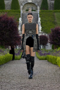 Female model in a metallic, armor-inspired tunic with belt and gauntlets walks a gravel garden path wearing knee-high boots.