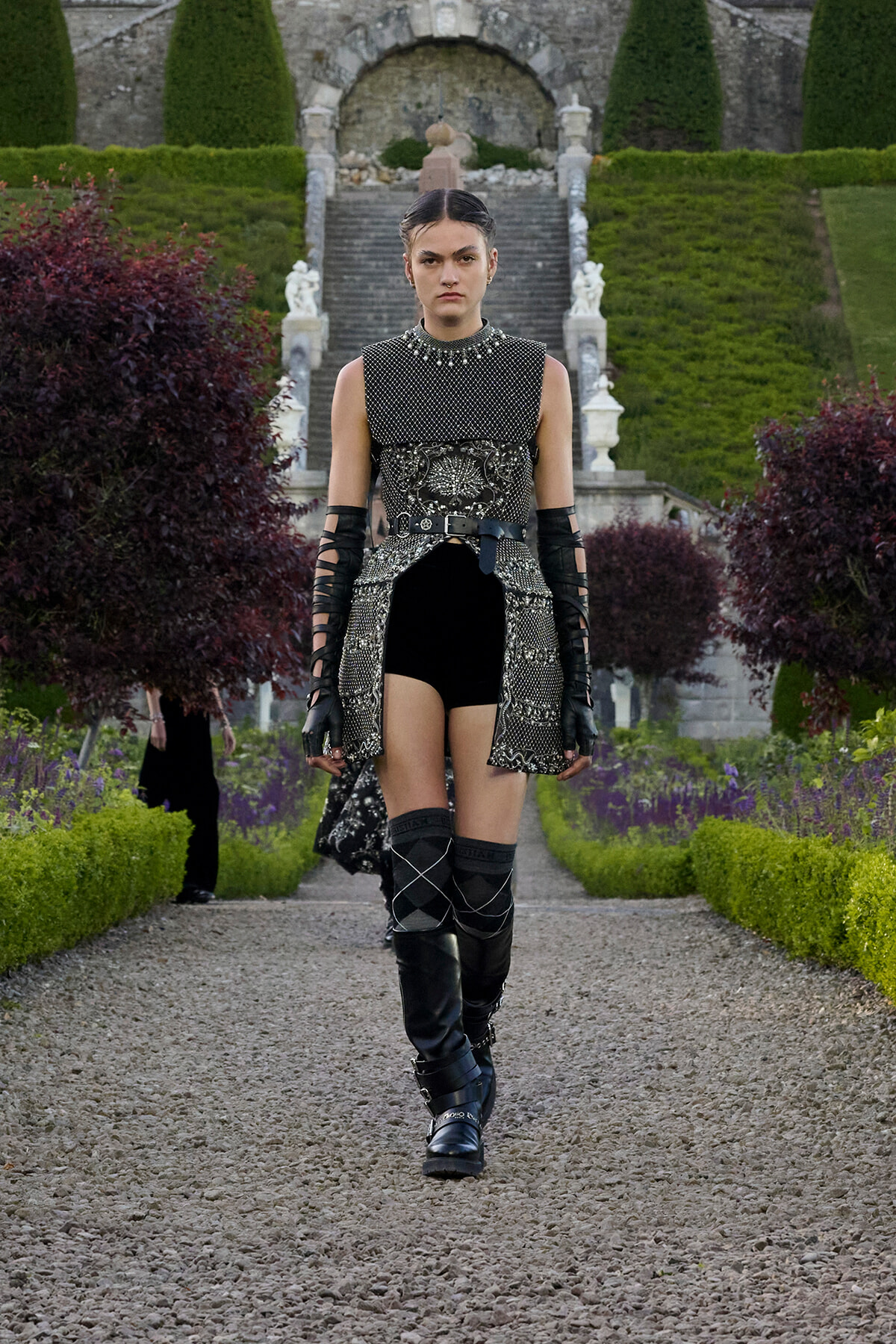 Female model in a metallic, armor-inspired tunic with belt and gauntlets walks a gravel garden path wearing knee-high boots.