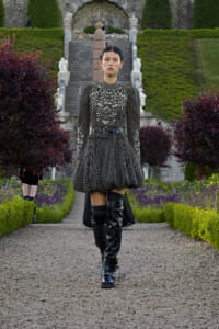 Model wearing a metallic, beaded dress with puff shoulders and a wide belt, walking along a gravel garden path in a formal landscape.