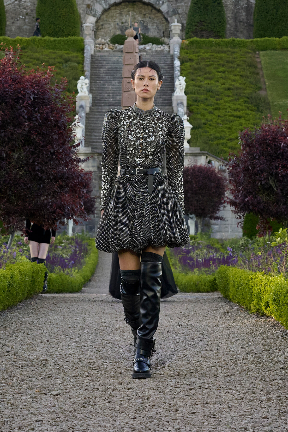 Model wearing a metallic, beaded dress with puff shoulders and a wide belt, walking along a gravel garden path in a formal landscape.