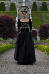 Model in a dramatic black gown with ornate embroidery, standing on a gravel path in a formal garden with trimmed hedges and stone stairway in the background.