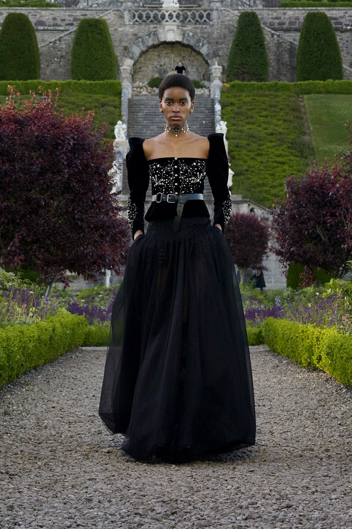 Model in a dramatic black gown with ornate embroidery, standing on a gravel path in a formal garden with trimmed hedges and stone stairway in the background.