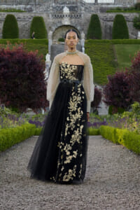 Model in a black gown with gold floral embroidery and sheer beaded cape, standing in a formal garden.