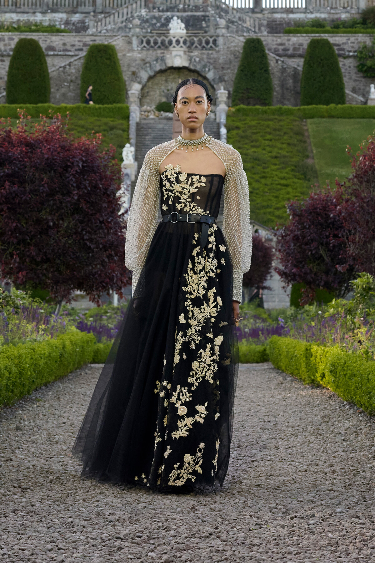Model in a black gown with gold floral embroidery and sheer beaded cape, standing in a formal garden.