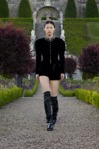 Model walking down a gravel path in a black velvet outfit with tall boots and a jeweled collar in a formal garden setting.