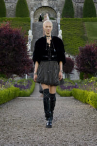 Model walking down a gravel garden path in a formal estate, wearing a black velvet top, dotted skirt, and tall boots.