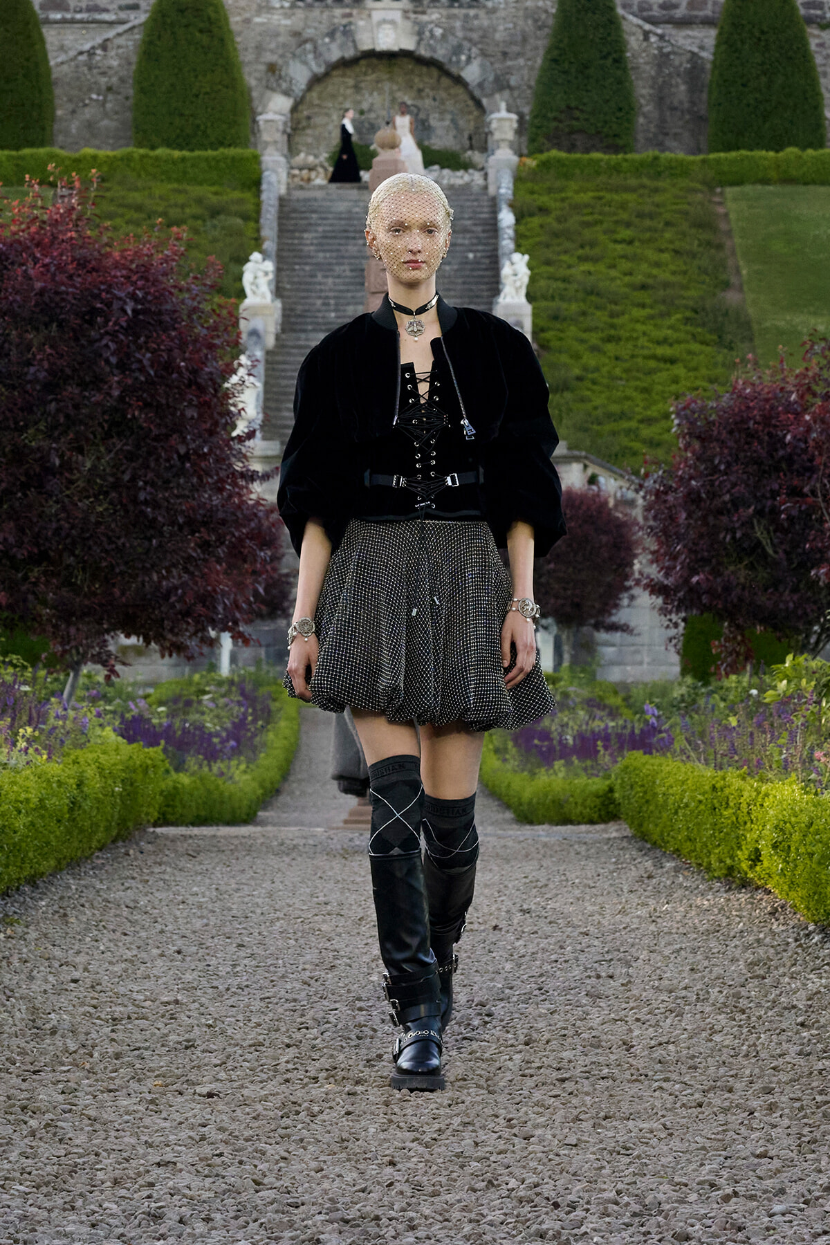 Model walking down a gravel garden path in a formal estate, wearing a black velvet top, dotted skirt, and tall boots.