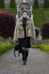 Model wearing a thick fur coat and black outfit walks down a gravel garden path with manicured hedges and stone stairs in the background.