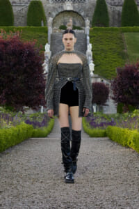 Fashion model walking toward the camera in a studded black outfit with cape sleeves, belt, and knee-high boots in a formal garden path.