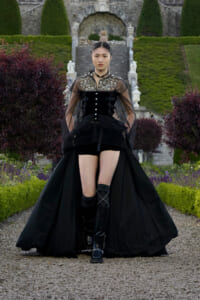 Person wearing a black Victorian-inspired gown with sheer sleeves standing in a formal garden near a stone stairway and archway behind them.