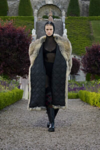 Model walking down a gravel garden path in a black sheer outfit under a long fur-trimmed quilted coat with a hooded silhouette.