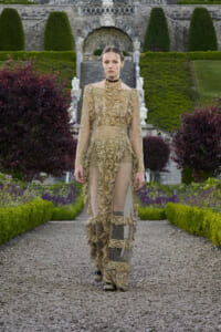 Model in an ornate gold lace gown walking down a gravel garden path with manicured hedges and stone stairway in the background