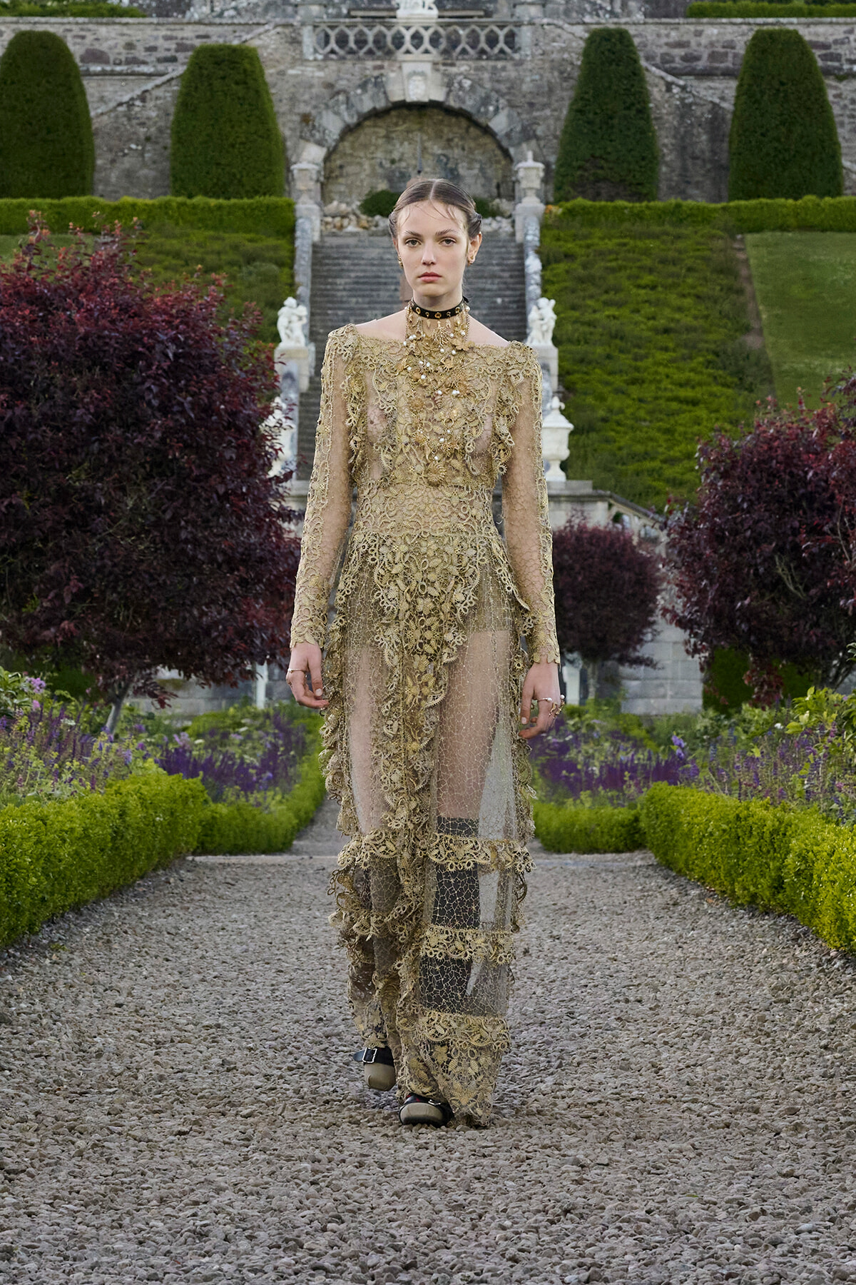 Model in an ornate gold lace gown walking down a gravel garden path with manicured hedges and stone stairway in the background