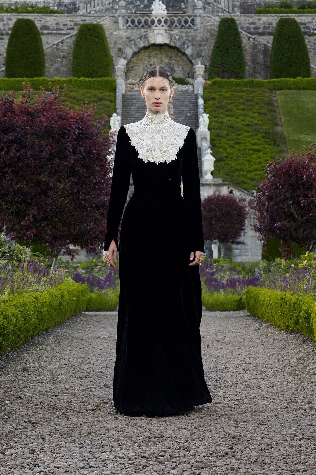 Woman in a long black velvet gown with a high white lace collar standing on a gravel garden path.