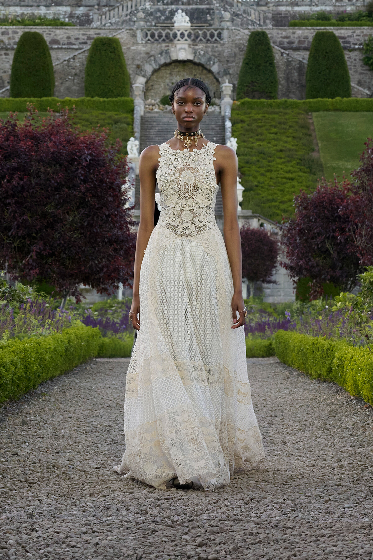 Model in ivory lace gown standing on a gravel path in a formal garden with stone stairs behind.