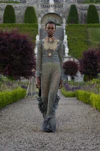 Model wearing an ornate lace jumpsuit with gold embroidery walks along a gravel garden path, stone stairs and hedges in the background.
