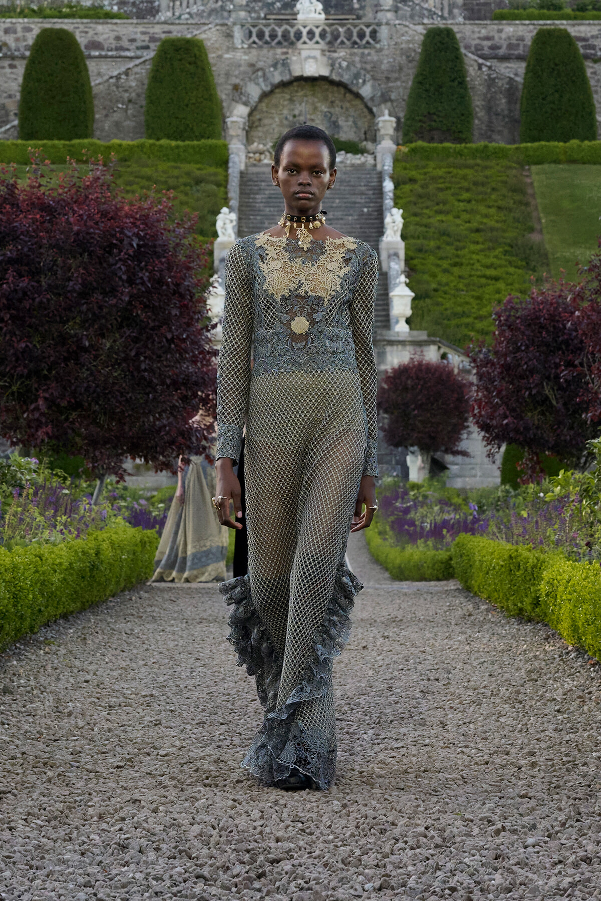 Model wearing an ornate lace jumpsuit with gold embroidery walks along a gravel garden path, stone stairs and hedges in the background.