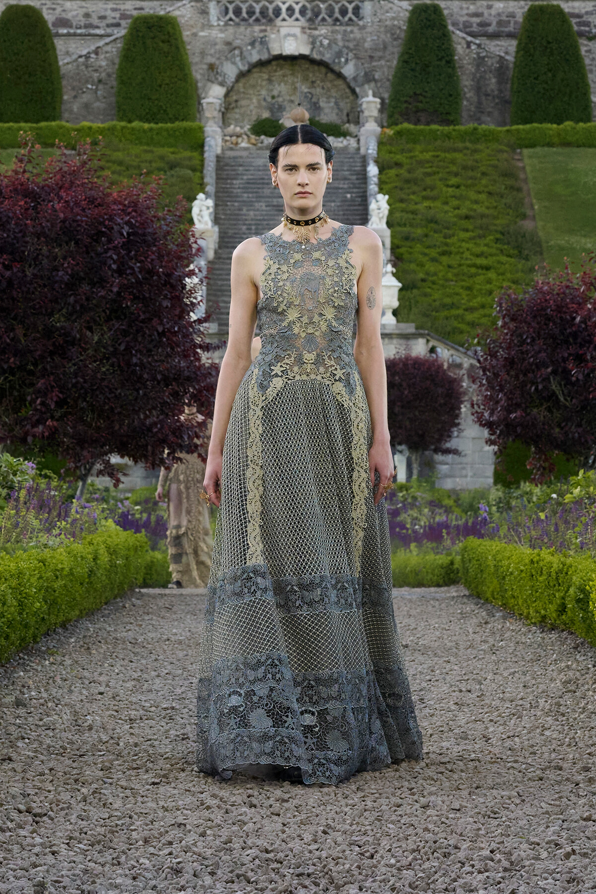 Model wearing an intricate gray lace gown standing on a gravel garden path with manicured hedges and stone stairs behind her.