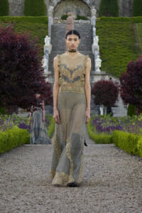 Model wearing an ornate beige lace dress with sheer skirt walking down a gravel garden path, with manicured hedges and stone steps in the background.