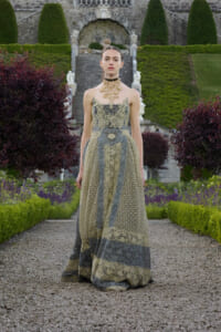 Fashion model wearing an intricate gold-and-gray lace gown stands on a gravel path in a formal garden with trimmed hedges and stone steps behind her.