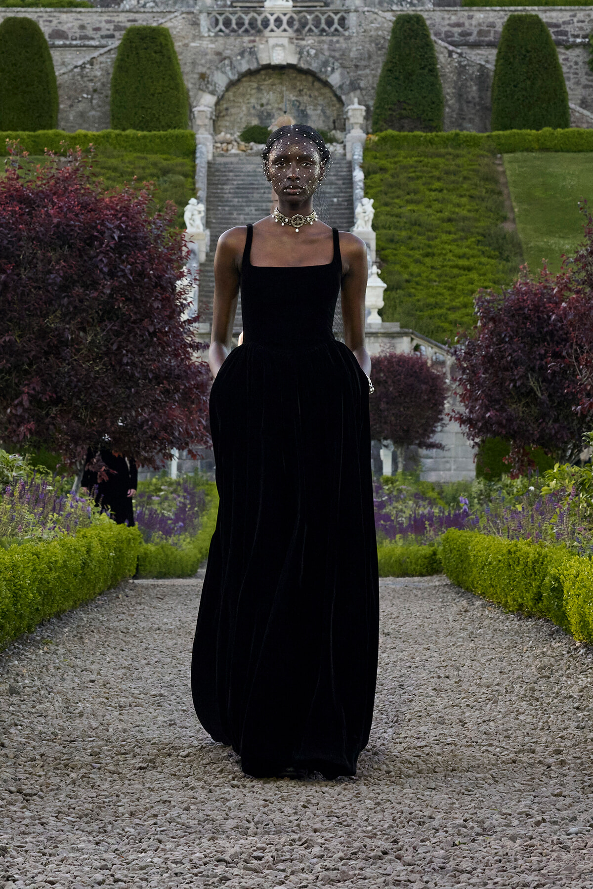 Elegant dark-skinned model in a long black velvet gown with thin straps walks down a gravel path in a formal garden, wearing a beaded veil and choker.