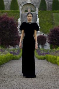 Model in a floor-length black gown standing on a gravel path in a formal garden, wearing a pearl necklace and bracelets with a sheer dotted veil over her face.