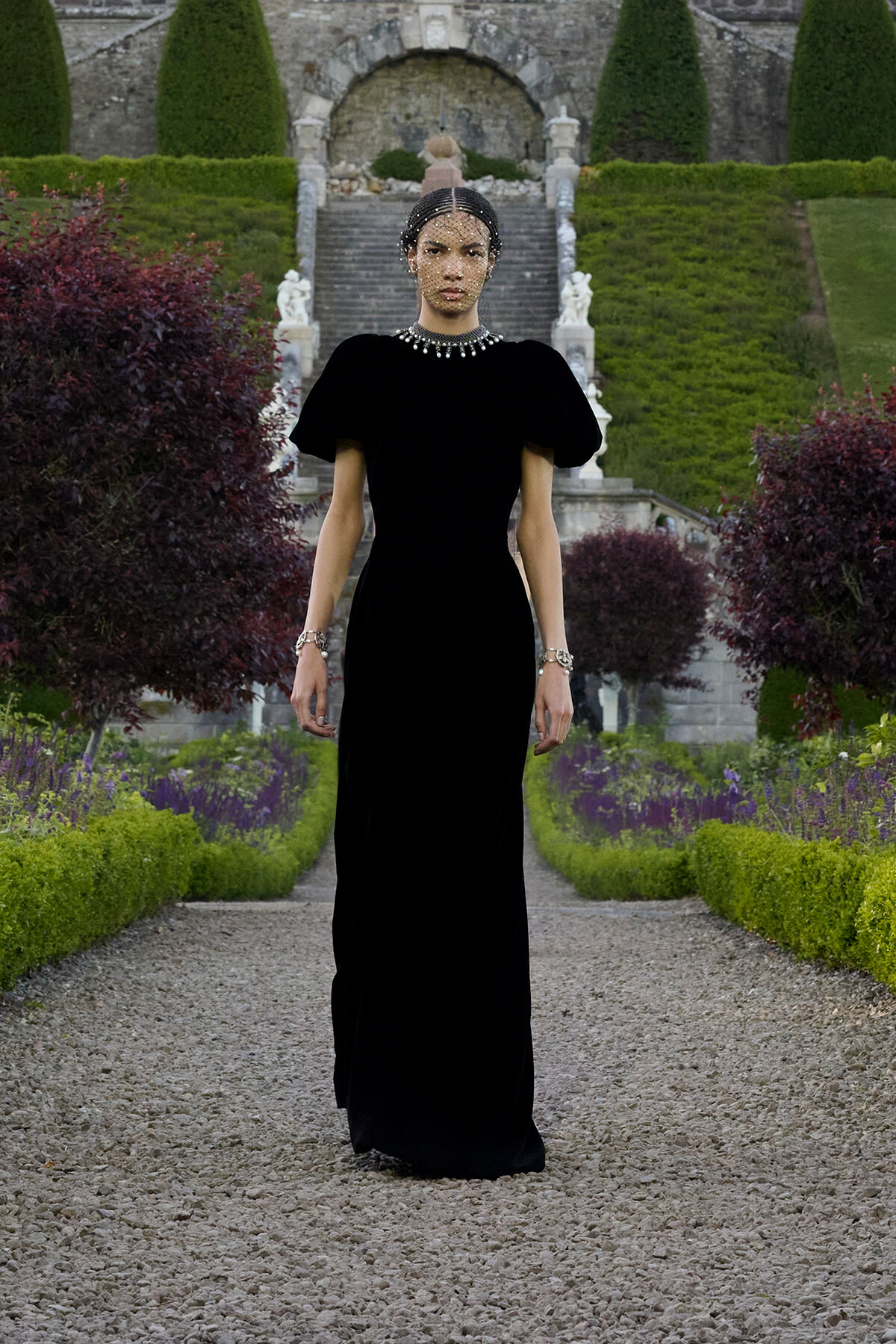 Model in a floor-length black gown standing on a gravel path in a formal garden, wearing a pearl necklace and bracelets with a sheer dotted veil over her face.