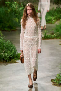 Model walking a runway in a long ivory lace dress with floral appliques, carrying a brown handbag.