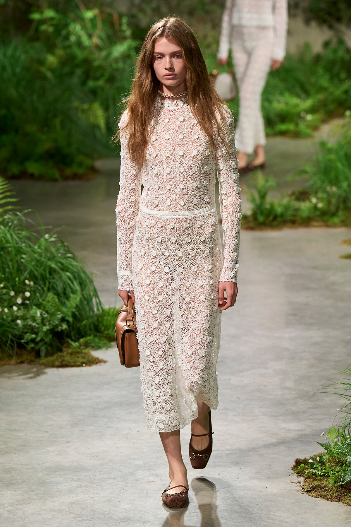 Model walking a runway in a long ivory lace dress with floral appliques, carrying a brown handbag.