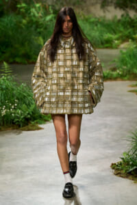 Model walking on a garden runway in a gold metallic grid-pattern poncho dress, paired with white socks and black loafers, pendant necklace visible.