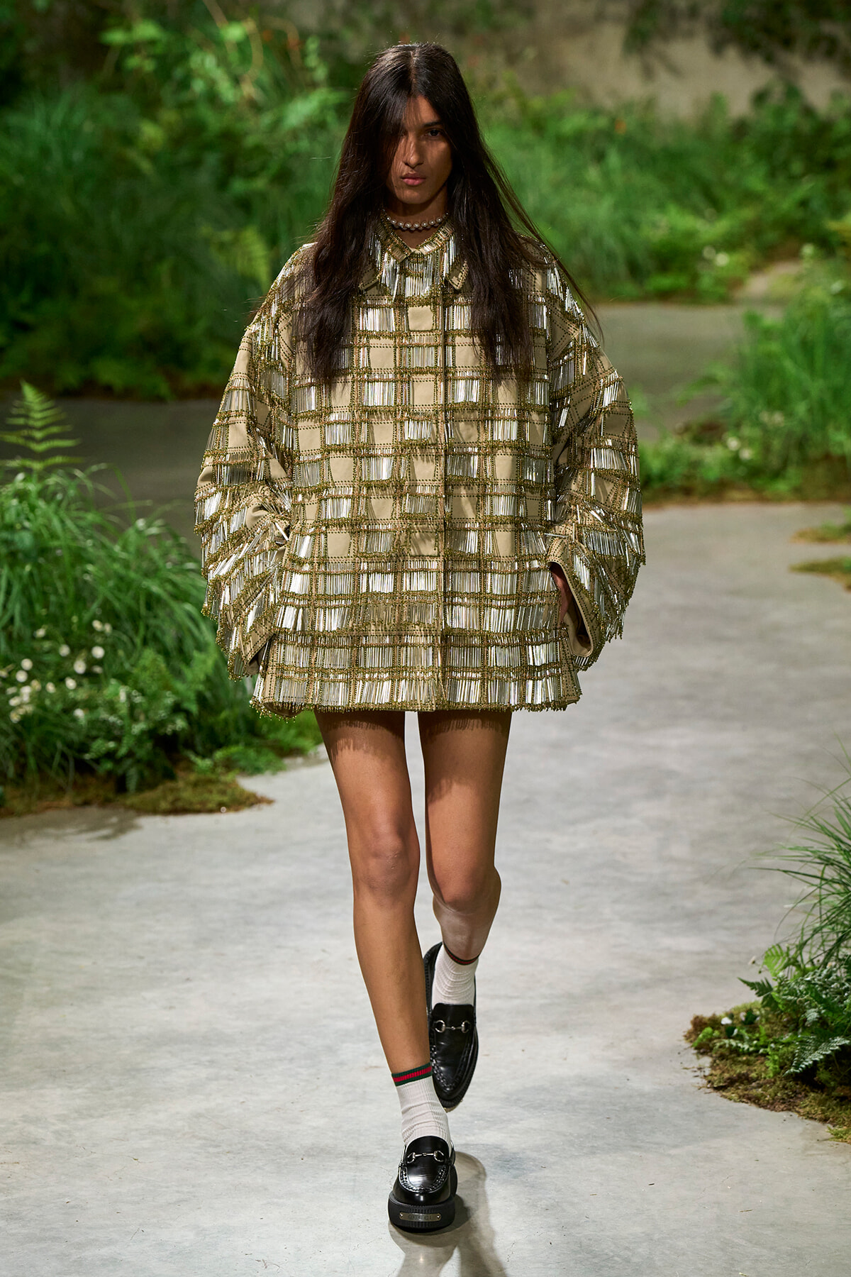 Model walking on a garden runway in a gold metallic grid-pattern poncho dress, paired with white socks and black loafers, pendant necklace visible.