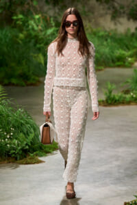 Woman walking along a garden path in a white lace two-piece outfit, wearing sunglasses and carrying a tan-and-white handbag.