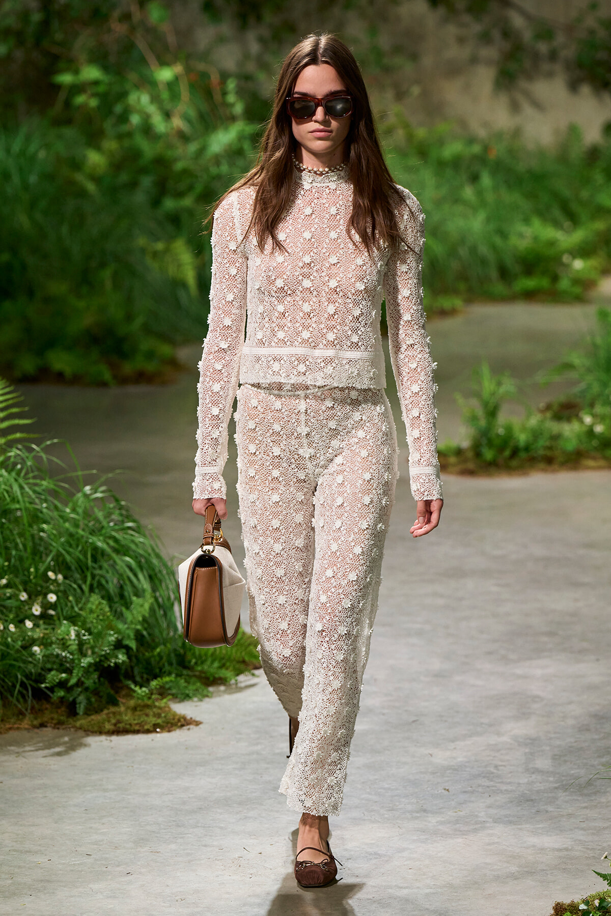 Woman walking along a garden path in a white lace two-piece outfit, wearing sunglasses and carrying a tan-and-white handbag.