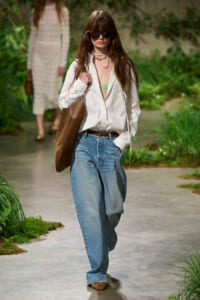 Model walking a garden runway in an oversized white shirt over a green lace bralette, loose blue jeans, brown belt, large brown tote, and sunglasses with layered necklaces.