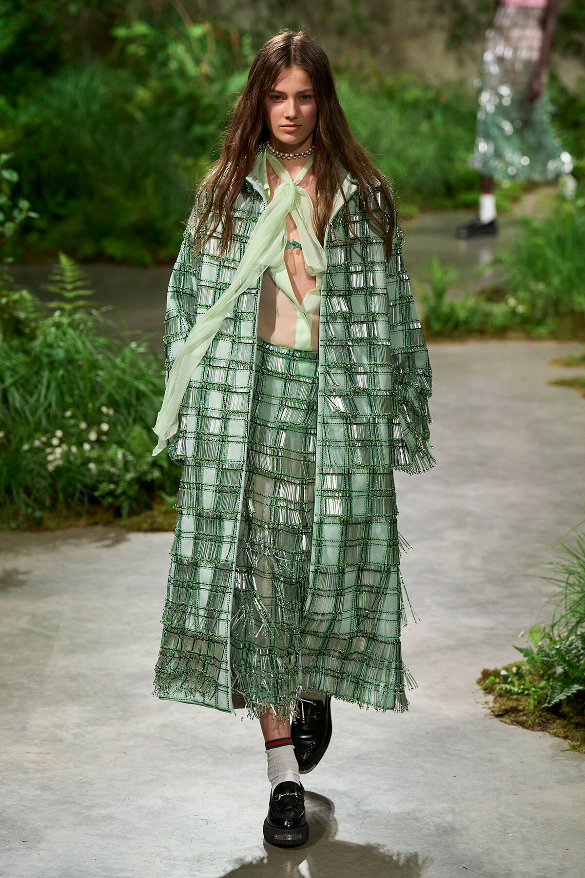 Model on an outdoor runway wearing a green plaid fringe coat over a mint blouse; black shoes and white socks visible.