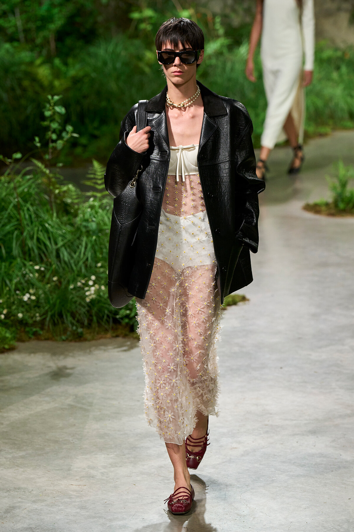 Model walks on a garden runway wearing an oversized black leather coat over a sheer pink-and-white floral midi dress, with a pearl necklace and dark sunglasses.