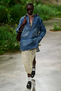 Model walking outdoors in an oversized denim shirt, daisy-print skirt, sunglasses, and loafers, carrying a brown bag.