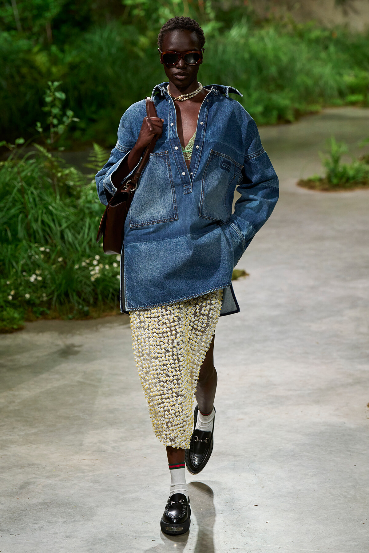 Model walking outdoors in an oversized denim shirt, daisy-print skirt, sunglasses, and loafers, carrying a brown bag.