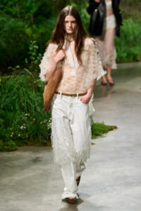 Female model walking a runway in a sheer pink embroidered blouse, white fringe trousers, brown belt and tan shoulder bag outdoors.