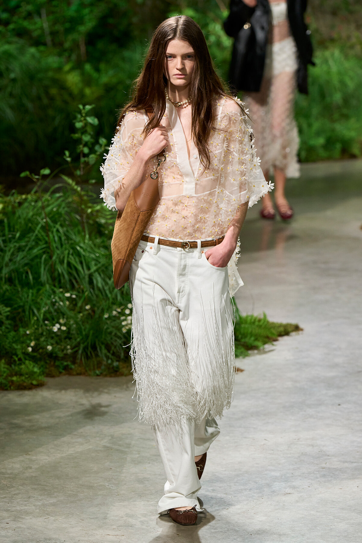 Female model walking a runway in a sheer pink embroidered blouse, white fringe trousers, brown belt and tan shoulder bag outdoors.