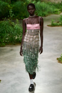 Black female model walking a garden runway in a pink satin bustier and a long, tiered beaded fringe skirt with green tones, paired with white socks and black loafers.