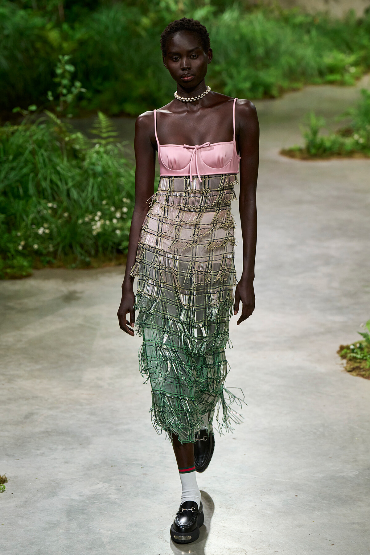 Black female model walking a garden runway in a pink satin bustier and a long, tiered beaded fringe skirt with green tones, paired with white socks and black loafers.