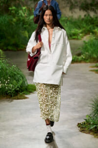 Model walking a garden runway in an oversized white shirt, daisy-print midi skirt, and burgundy handbag.